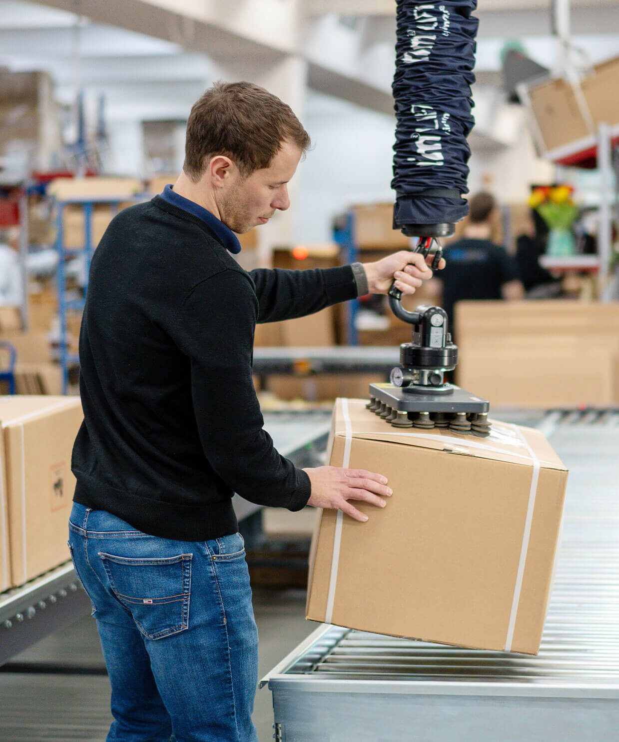 Ein Mann hebt ein schweres Paket mit einem modernen Vakuum Heber im Fullfilment Lager der Versandhelden